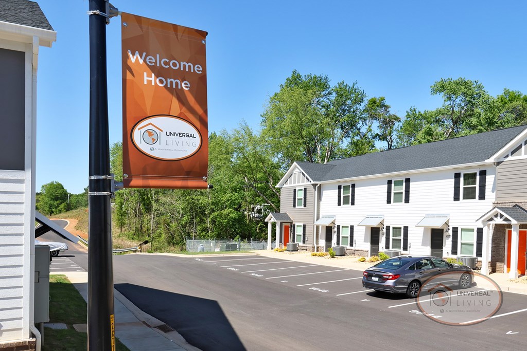 An orange flag reading "Welcome Home" stands before a road leading into the apartment home community.