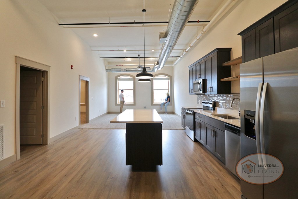 An empty apartment unit with light colored carpets, white walls, and a kitchen with dark cabinets and stainless steel appliances.