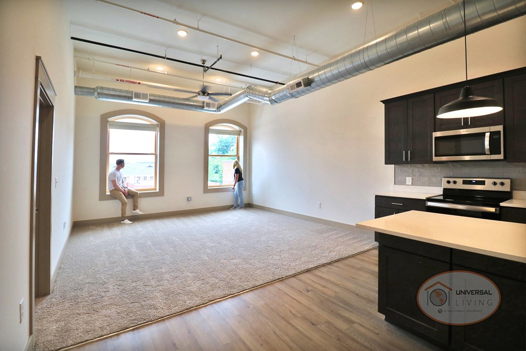 An empty apartment unit with light colored carpets, white walls, and a kitchen with dark cabinets and stainless steel appliances.