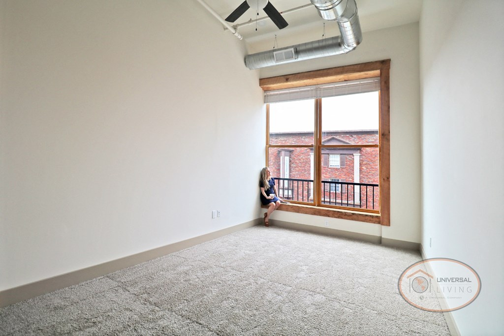 A woman sitting in the windowsill of a large window in a living room with white walls, carpet and a ceiling fan.