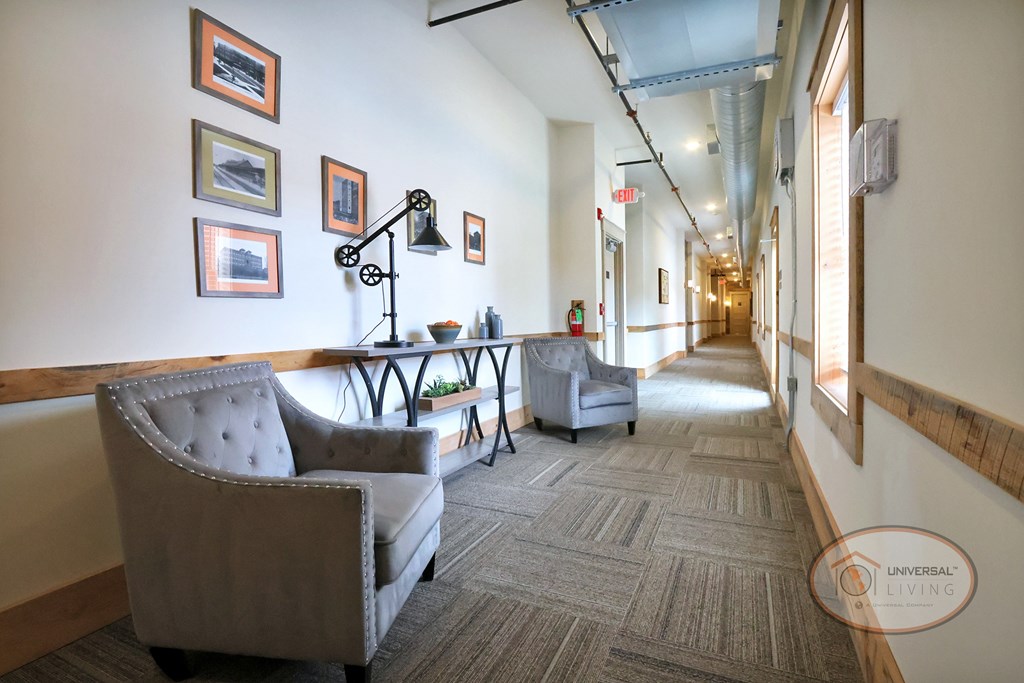 A hallway with a sitting area complete with table, lamp, two grey armchairs, and artwork.