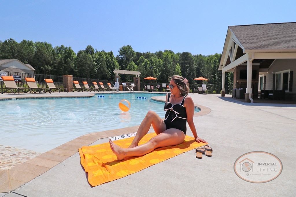 A woman in a black swimsuit is sitting on a yellow towel by a pool with a row of lounge chairs in the background.