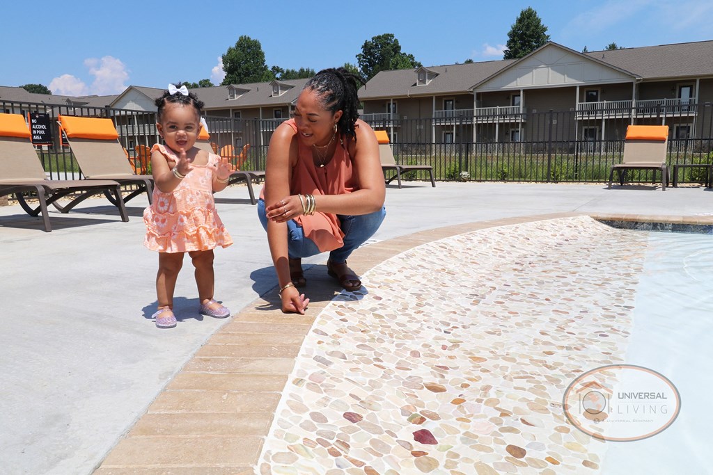 A woman and a little girl are playing by a pool in front of a row of lounge chairs.