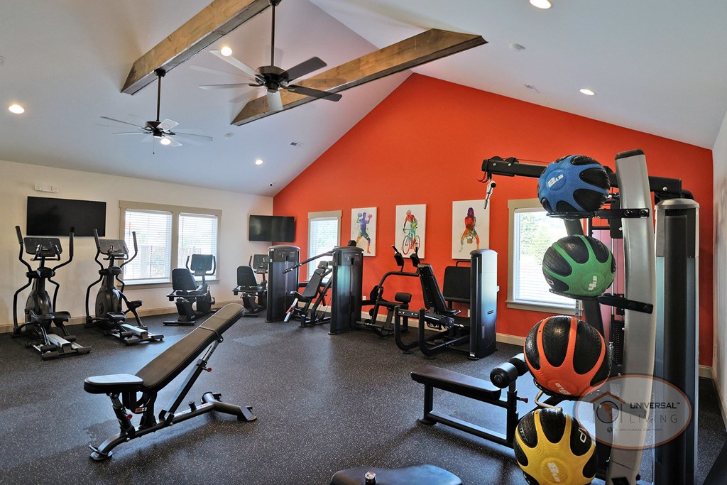 A fitness center with an orange accent wall, large windows, flat screen TVs, with weight training and cardio equipment.