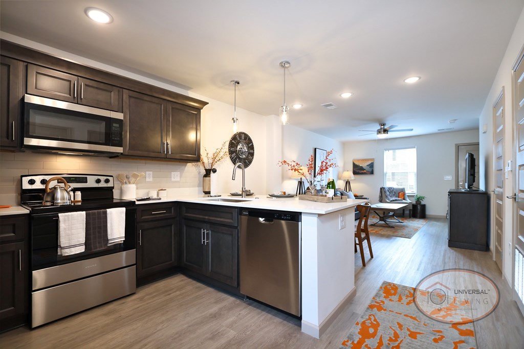 A modern kitchen with hardwood vinyl flooring, dark wood cabinets and stainless steel appliances. The living room can be seen behind the ktichen.