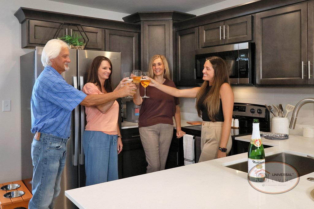 Four people toasting with drinks in a kitchen with stainless steel appliances and dark cabinets..