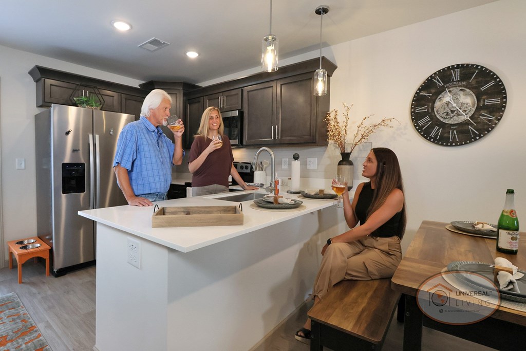A man and woman sip wine in the kitchen while another woman sits at a nearby dining table and chats with them.