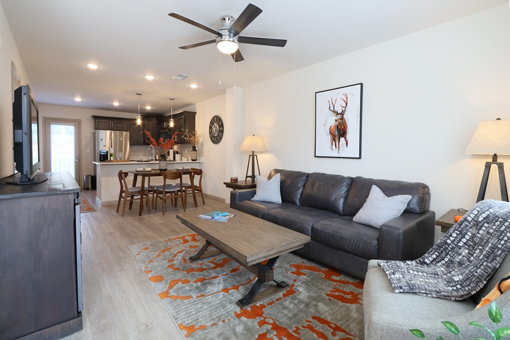 A fully furnished and spacious living room with hardwood vinyl flooring, white walls, and a ceiling fan.