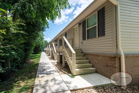 The front of a row of tan apartment buildings with a sidewalk leading to stairs.