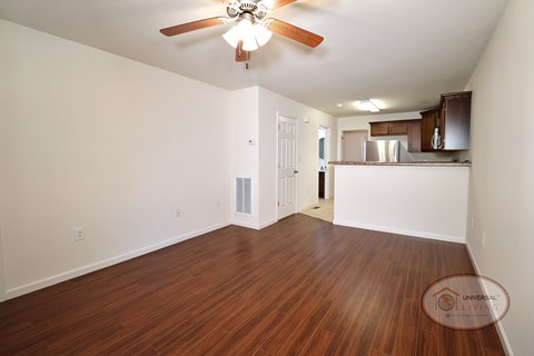 The living room with vinyl hardwood flooring, white walls, and a ceiling fan with a bar and kitchen in the background..