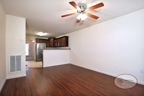 The living room with vinyl hardwood flooring, white walls, and a ceiling fan with a bar and kitchen in the background..