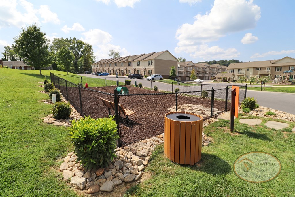 A fenced in dog park with obstacles, a water fountain for pets, and apartments in the background.