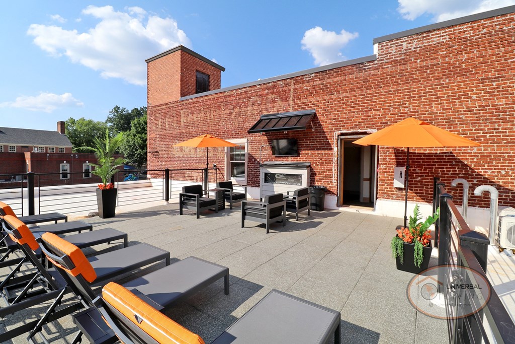 A fenced in, rooftop patio, complete with soft surface for noise cancellation, a fire place and flat screen TV, lounge chairs, tables, and umbrellas.