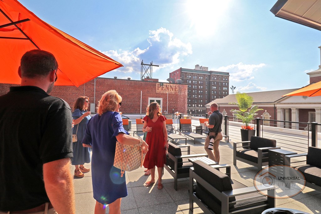A group of happy people relaxing on the rooftop patio with drinks. The sun shines brightly in the background, and the tops of historical buildings can be seen.