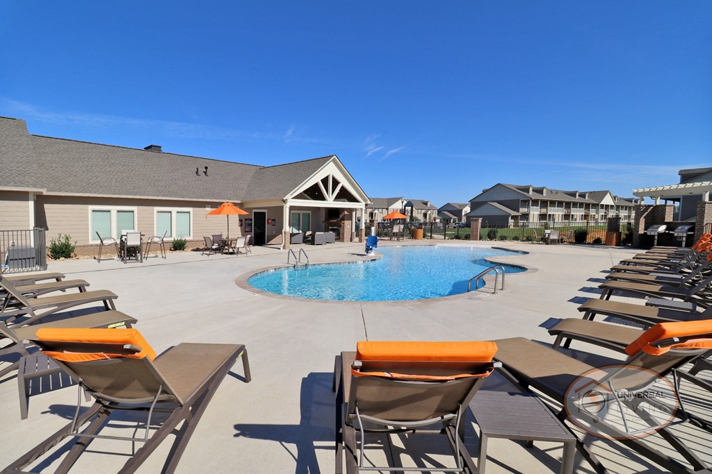 A large pool surrounded by lounge chairs with orange pillows and a clubhouse in the background.