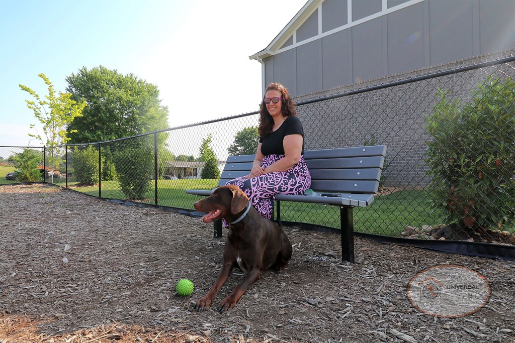 A woman sits on a bench with a her dog inside a fenced in dog park.