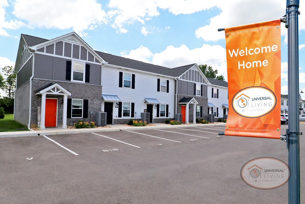 An image of a white and grey apartment home with orange doors and a flag that reads "Welcome Home."
