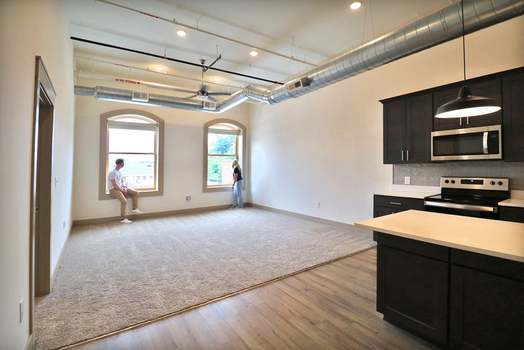 An empty, carpeted, living room with a man sitting in a window and a woman looking out of the other window.