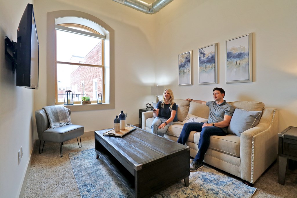 Two people sitting on a couch in a living room with white walls, a large window, and carpet.