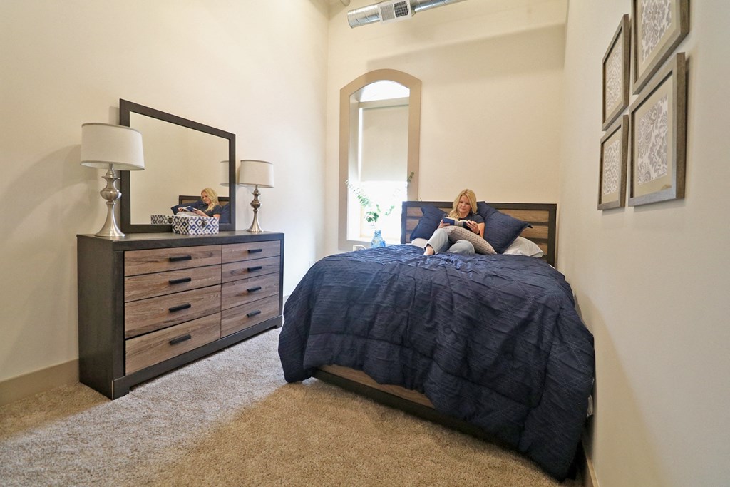 A woman sitting on a bed reading a book in a bedroom with a window and carpet.