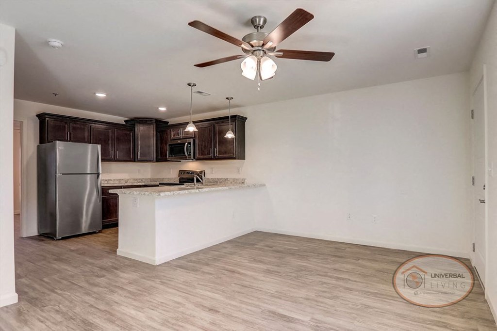 An empty living room with a kitchen and a ceiling fan.