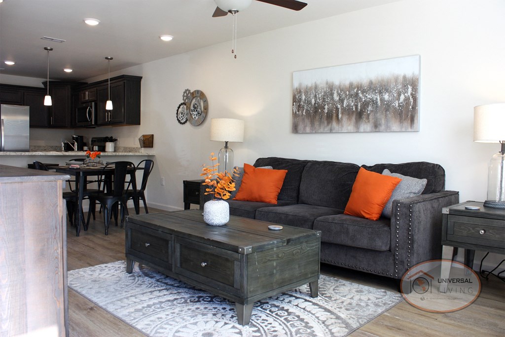 A living room with white walls, dark hardwood style vinyl flooring, and a kitchen visible in the background.