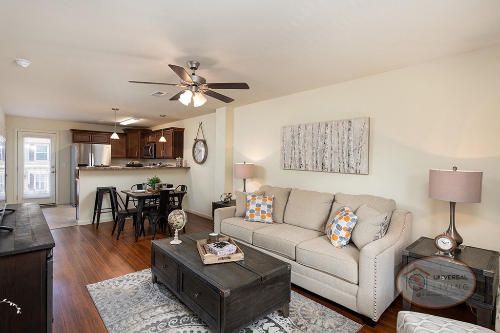 An open living area with white walls, hardwood vinyl flooring, a kitchen with dark cabinets, and a door to the patio.