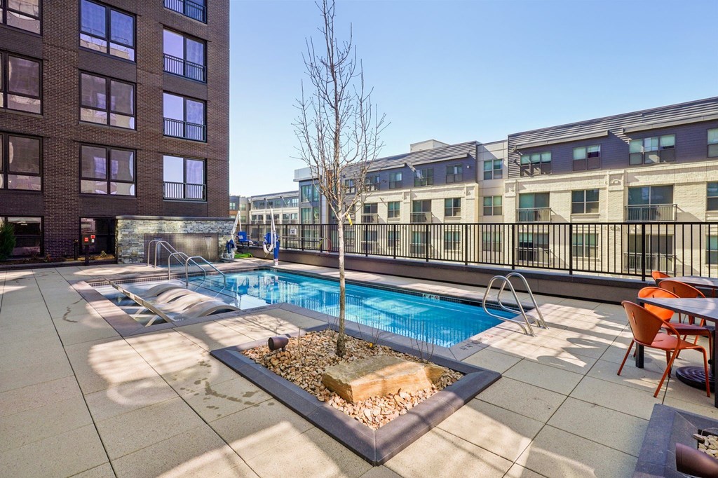 A small tree in a square planter is the centerpiece of a small pool surrounded by chairs and tables.