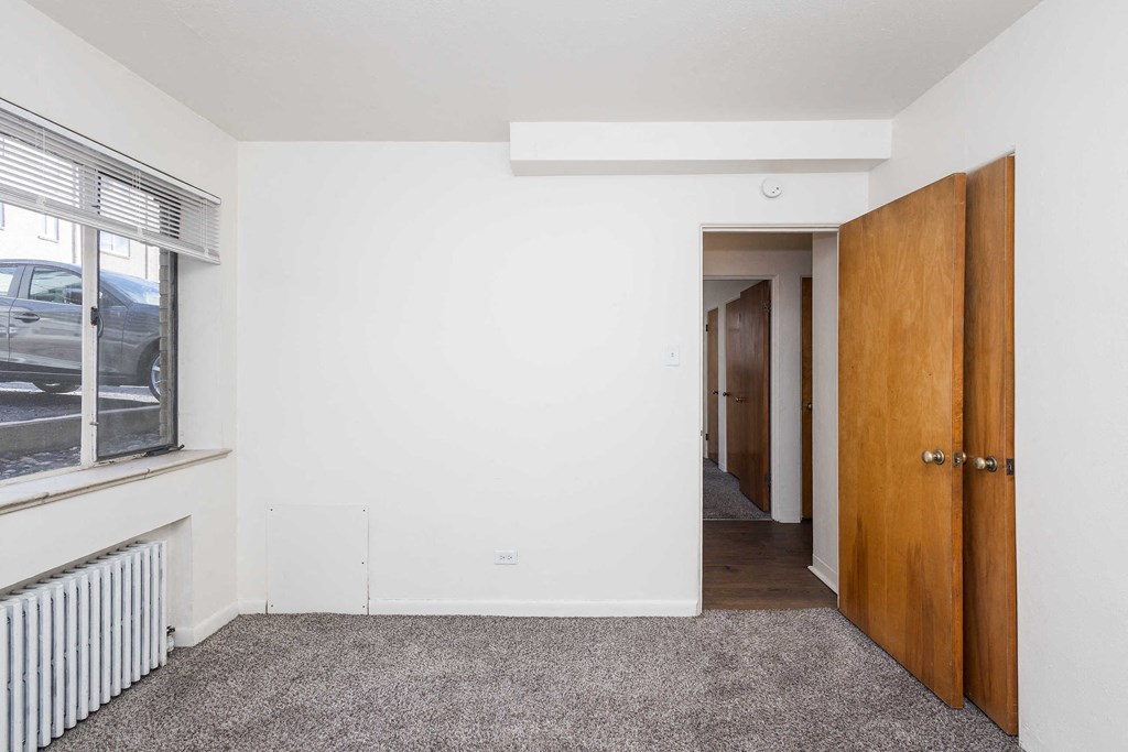 A room with a carpeted floor, a window with blinds, a radiator, and a wooden door.