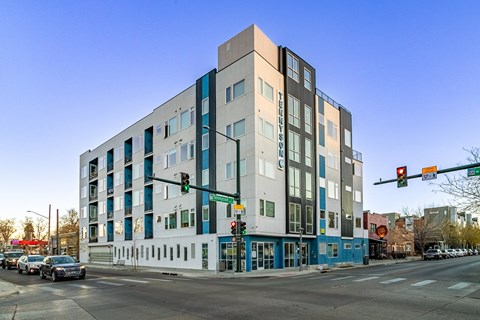 A modern multi-story building with a blue and white exterior is situated on a street corner with cars and traffic lights.