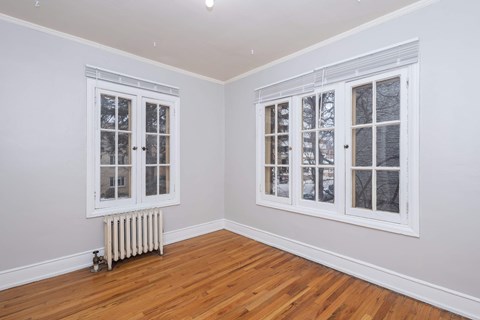 A room with a white wall and a white radiator with a window on the left and a door on the right.
