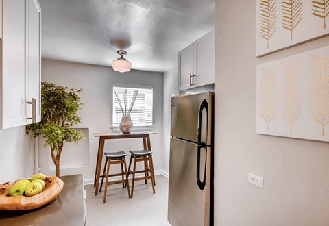 A kitchen with a refrigerator, a table with a vase and a bowl of fruit on it, and a plant.