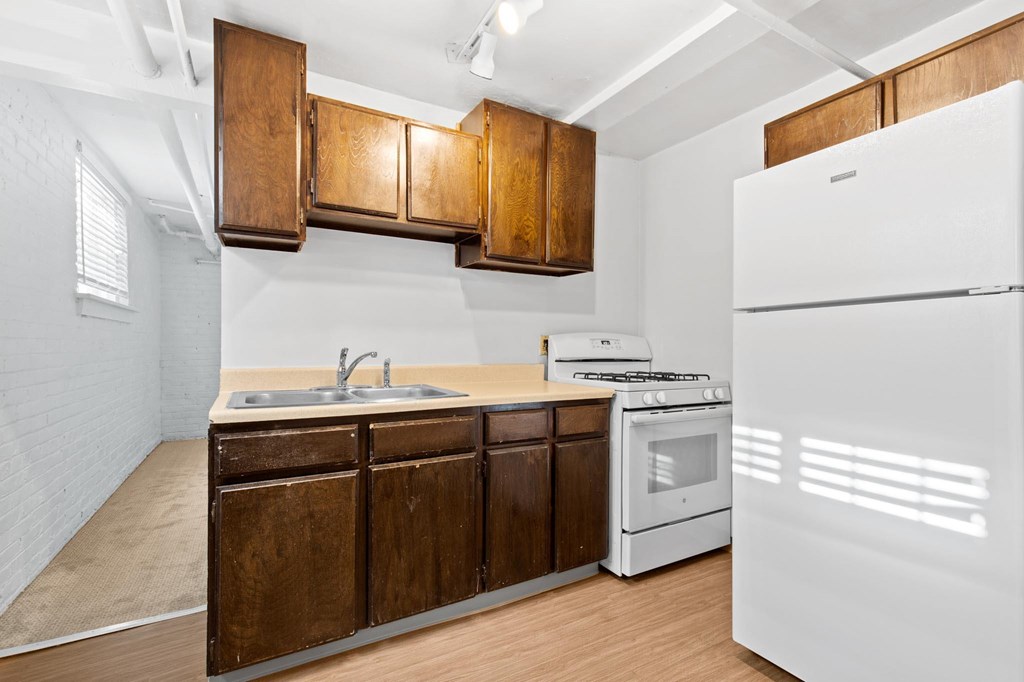A white refrigerator stands in a kitchen with wooden cabinets and a white sink.