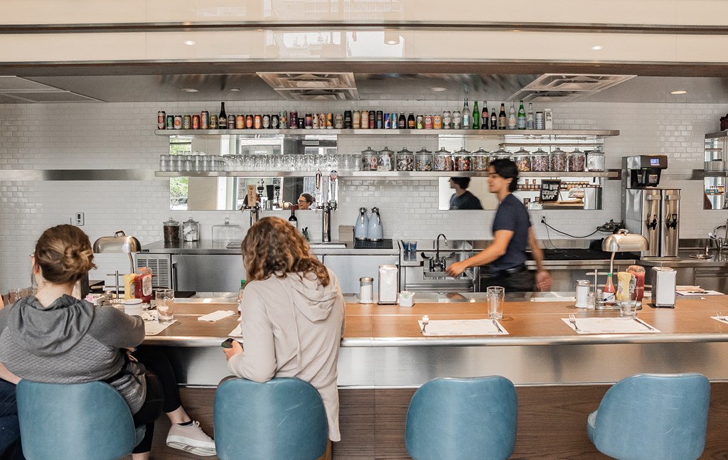two women sitting at a bar in a restaurant with a chef in a kitchen
