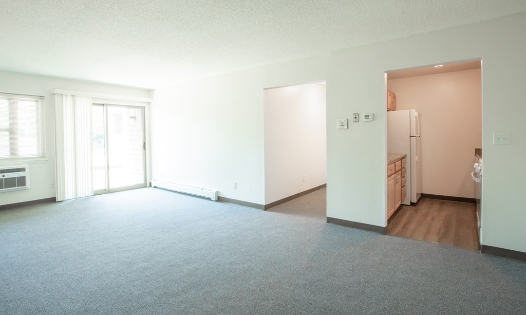 an empty living room with a refrigerator and a door to a kitchen
