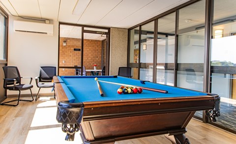 Billiards room with a pool table with a green cloth and white cue balls is in the foreground of a room with a brick wall and sliding glass doors.