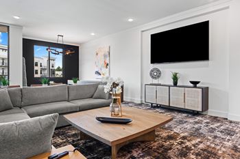A modern living room with a grey sofa, a wooden coffee table, and a large flat screen TV mounted on the wall at Views at Flowers Apartments, North Carolina