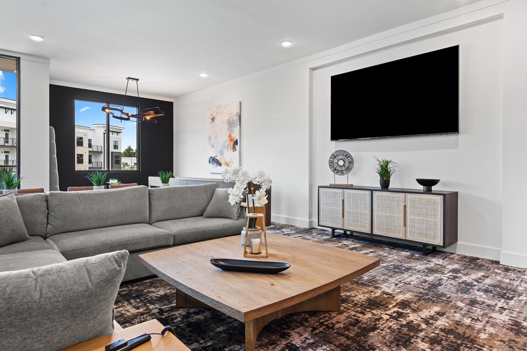 A modern living room with a grey sofa, a wooden coffee table, and a large flat screen TV mounted on the wall at Views at Flowers Apartments, Clayton