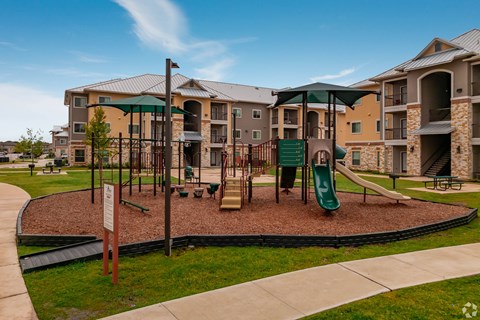 A playground area with a green slide and a brown sandbox in front of apartment buildings.