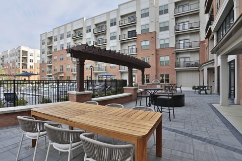 A wooden table with chairs is in the foreground of a patio area with a building in the background.