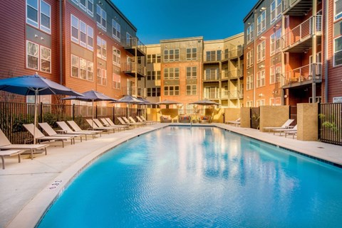 A large swimming pool surrounded by lounge chairs and umbrellas in front of a multi-story apartment building.