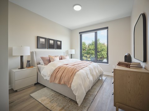A bedroom with a large bed, a nightstand with a lamp, a dresser, and a window with a view of trees.
