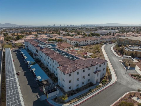 A large white building with red tile roofs is surrounded by a parking lot and a road.