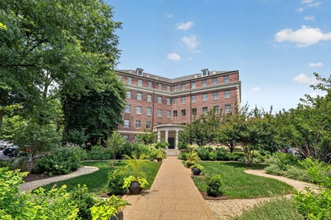 A large building with a red brick exterior is surrounded by a well-manicured garden with a gravel path leading to the entrance.