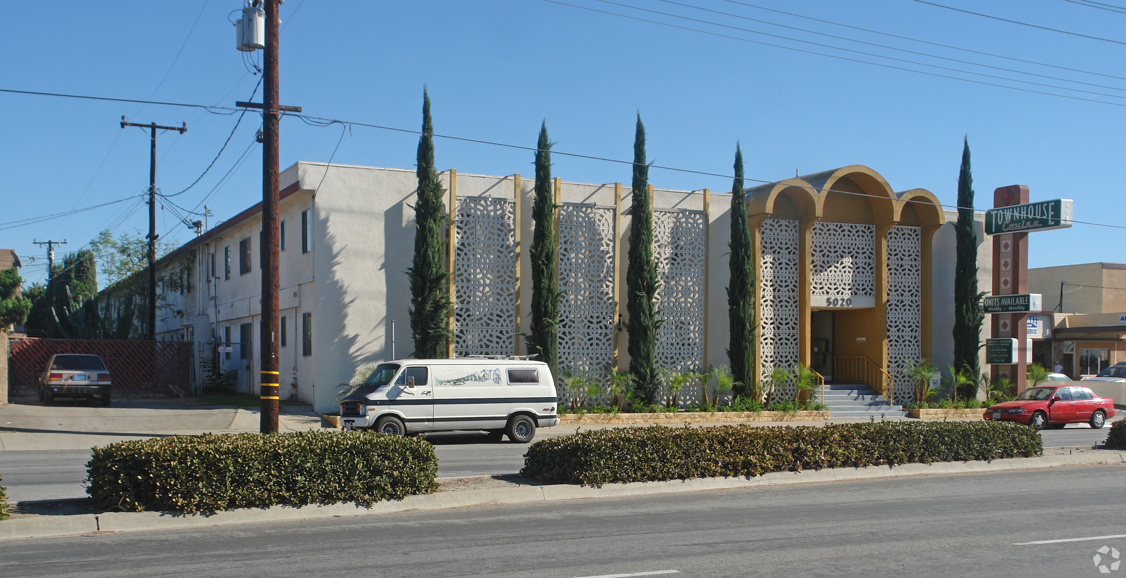 A white building with a yellow arched doorway is behind a row of green trees.