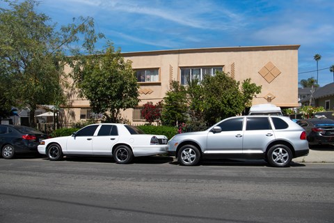 Two cars parked in front of a tan building with trees in the background.