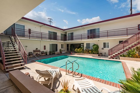 A pool surrounded by chairs and plants is in the foreground of a building with a red roof.