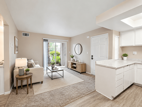 A well-lit, spacious living room with a white sofa, a wooden side table, and a large window.