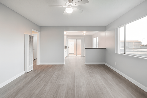 A spacious room with a ceiling fan and light, hardwood flooring, and a large window with blinds.