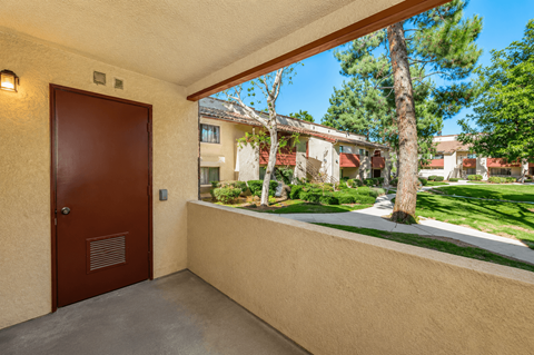 A brown door is on the left of a balcony with a view of a green lawn and trees.